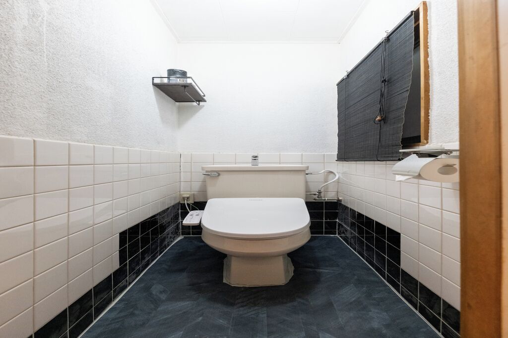Une petite salle de bain étroite, avec des toilettes, une étagère au-dessus des toilettes et une fenêtre sur le côté droit.