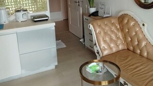 Moderni keittiö, jossa on ruostumattomasta teräksestä valmistettu liesituuletin, valkoinen työtaso ja kahvinkeitin. Olohuoneessa on beige sohva ja lasinen sohvapöytä.