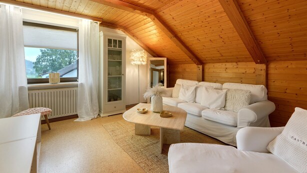 Een knus vertrek met een houten plafond, witte meubels en een groot raam met gordijnen.