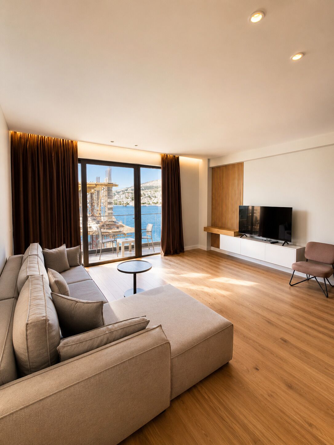 A modern living room with a large sofa, a small round table, a television, and a view of a cityscape through large windows.