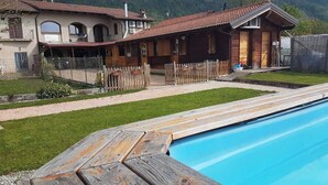 Un espace aménagé au bord de la piscine avec une terrasse en bois, une maison avec un toit en tuiles et une cour clôturée.