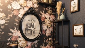Una habitación con papel tapiz floral, una fotografía blanco y negro enmarco, un estante pequeño con objetos decorativos y un soporte de metal negro.