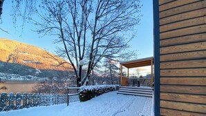 Een houten blokhut met een overdekt terras, een sneeuwbedekt dek en uitzicht op een meer en bergen.