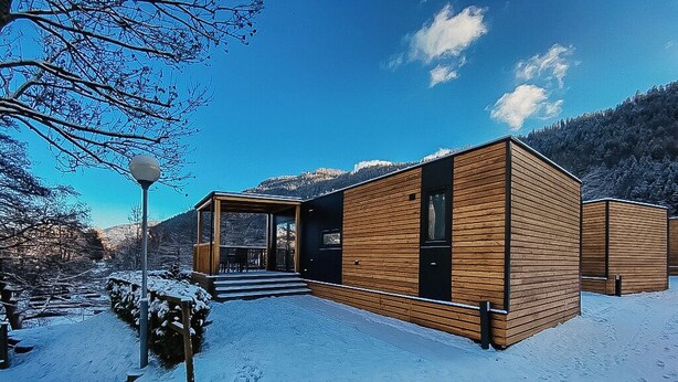 Een moderne houten chalet met een glazen ingang, gelegen in een besneeerd landschap met bergen op de achtergrond.