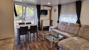 Een moderne woonkamer met een bank, een glazen salontafel, een eettafel met stoelen, een aan de muur bevestigde televisie en een uitzicht op de omgeving door grote ramen.