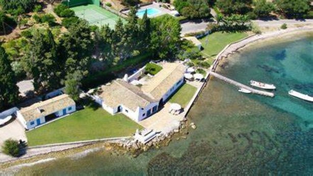 Vista aérea de uma propriedade costeira com piscina, um cais com barcos e área gramada.