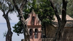 Um edifício rústico de pedra com um campanário, cercado por árvores e com vista para um corpo d'água.