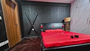 Une chambre avec un lit noir, une literie rouge, une porte en bois, un mur noir, un tapis rouge et une lampe.