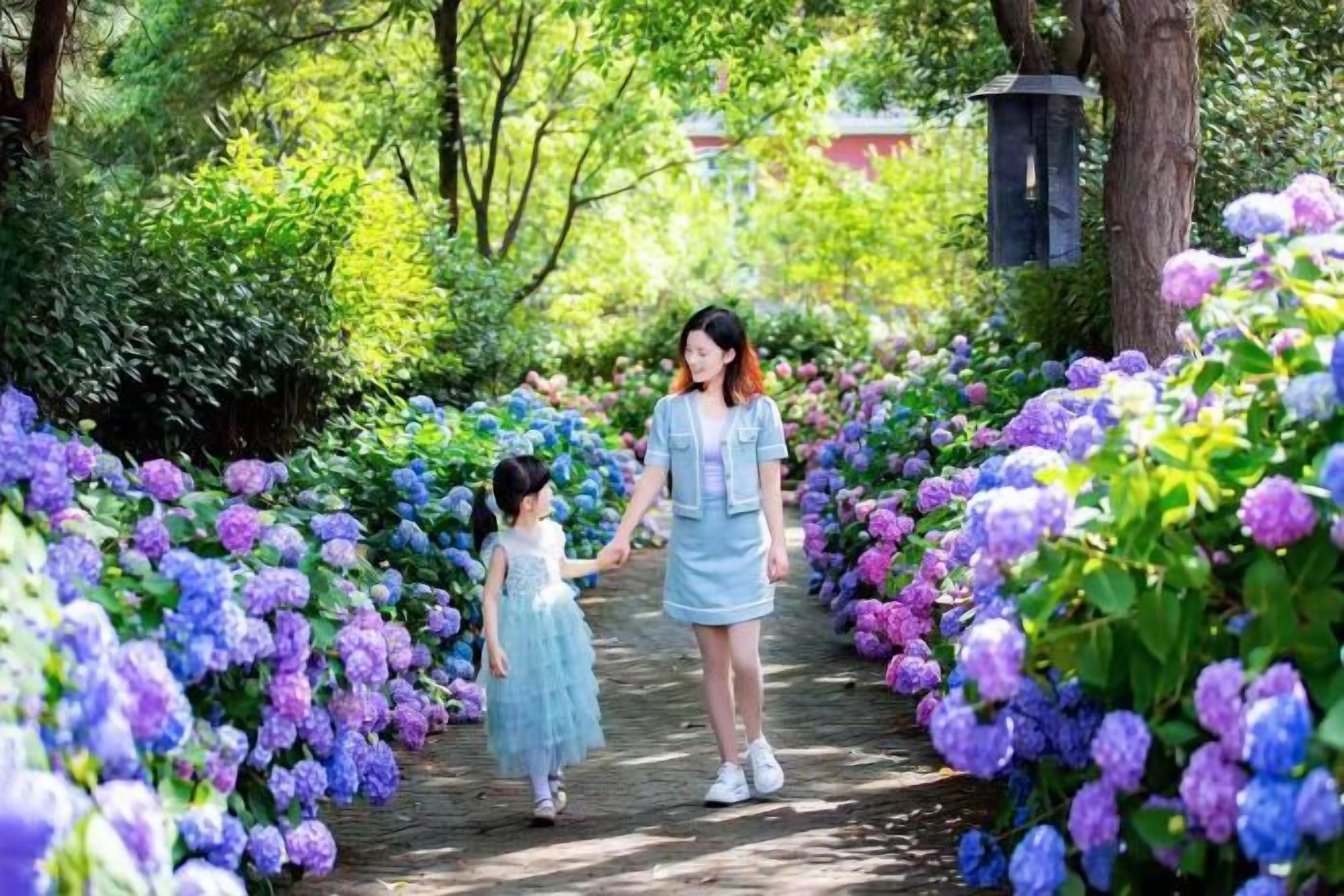 A pathway surrounded by vibrant flowers, a person and a child walking, and a lamp post in the background.