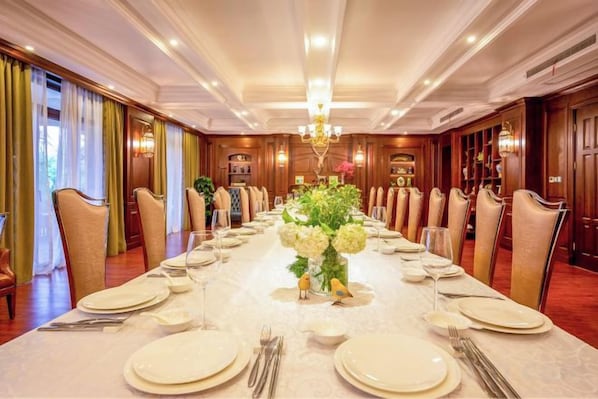 A long dining table set for a formal meal with chairs on both sides, a chandelier above, and a red carpeted floor.