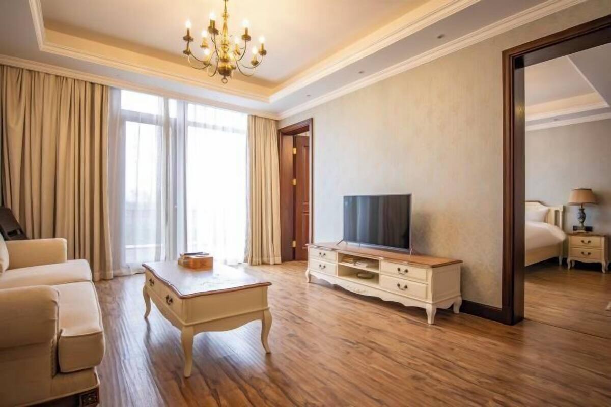 A hotel room with a sofa, a TV on a wooden stand, a bedside table, and a large mirror.