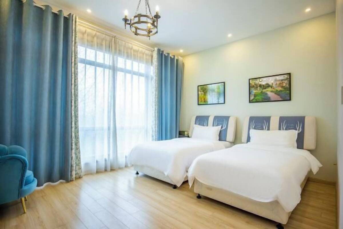 A hotel room with two beds, a chandelier, a blue chair, and two framed pictures on the wall.