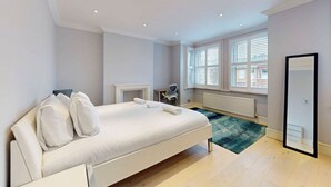 Une chambre moderne avec un grand lit, un petit bureau, un miroir et une vue sur l’extérieur grâce à de grandes fenêtres.