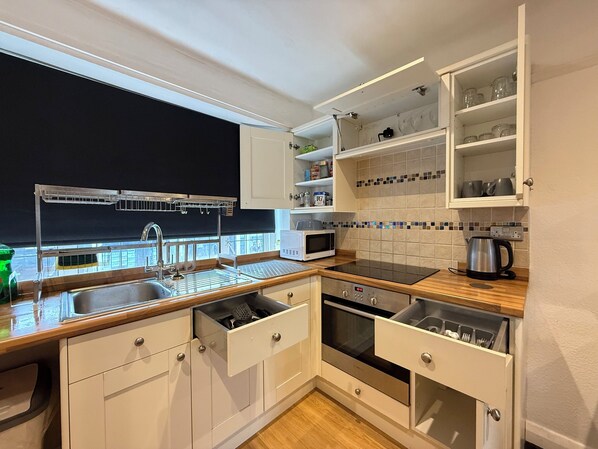 A modern kitchen with a built-in oven, microwave, and sink. It features white cabinetry, a wooden countertop, and a window overlooking a balcony.