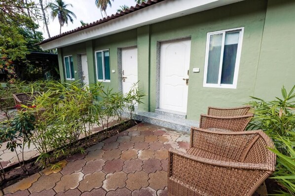 A row of green-walled rooms with white doors and windows, a tiled patio with wicker furniture, and lush greenery. - Oceanature Resort Alibaug (Alibag)
