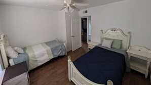 Une chambre avec deux lits simples et un lit plus grand, un ventilateur de plafond et une porte donnant sur une autre pièce.
