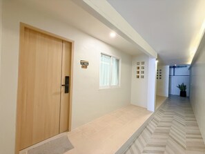 Un couloir avec une porte en bois, une fenêtre ornée d’un rideau blanc, un sol carrelé et une plante en pot.