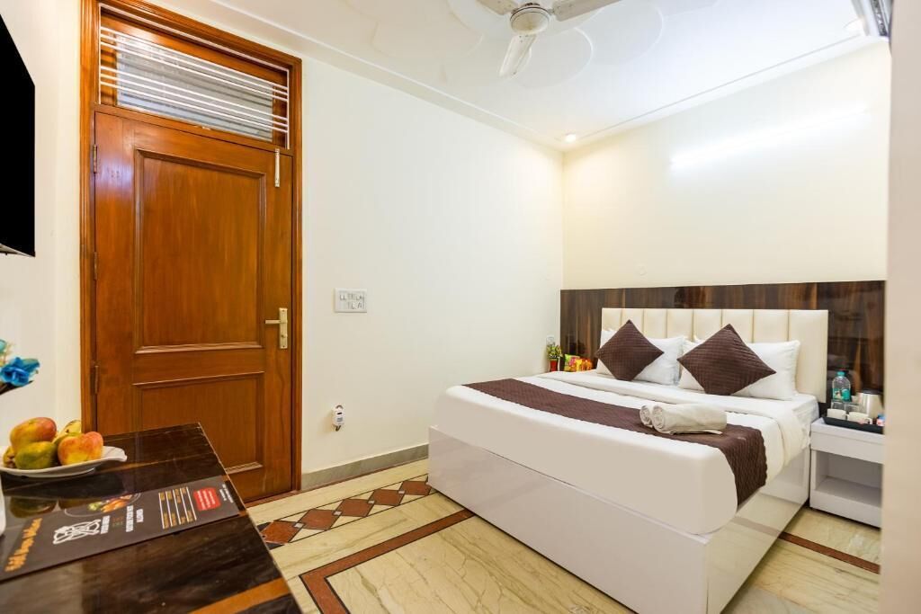 A hotel room with a wooden door, a bed with white and brown bedding, a bedside table with a bottle, and a patterned floor.