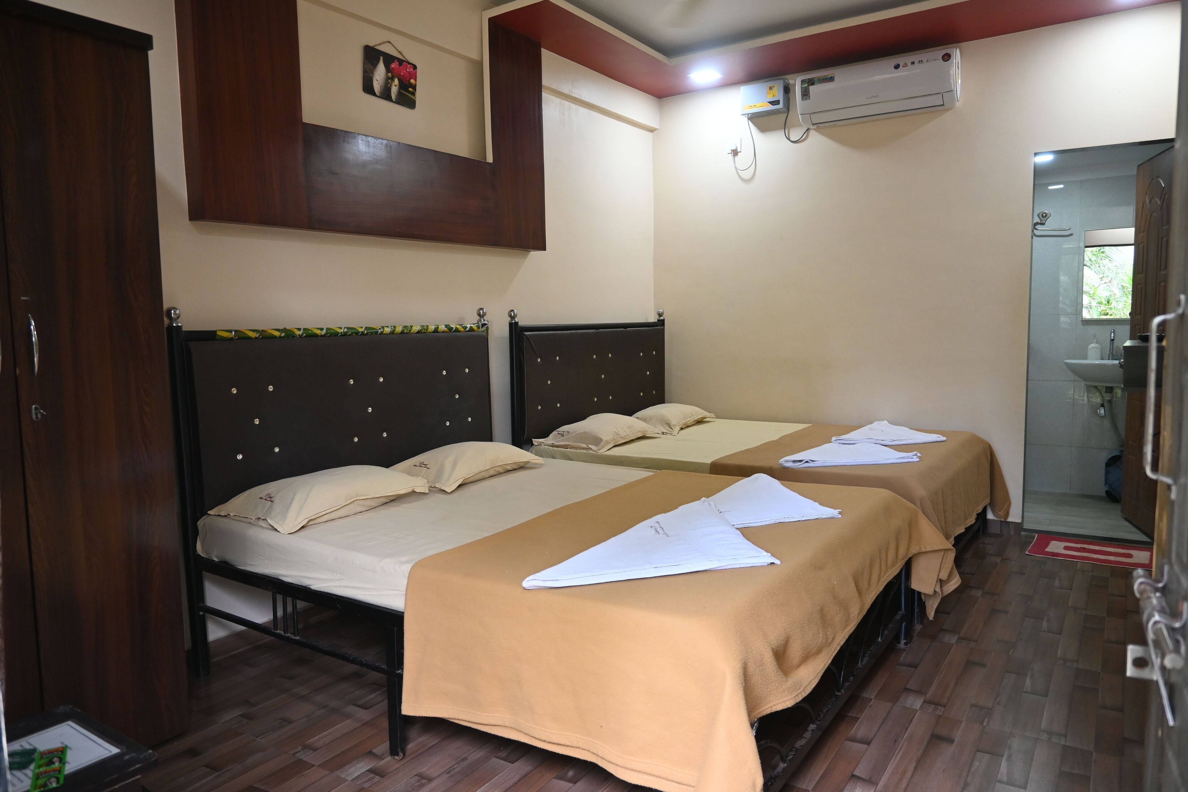 A hotel room with two beds, wooden flooring, a wardrobe, an air conditioning unit, and a bathroom visible through an open door.