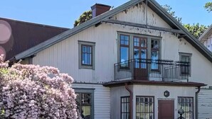 A two-story building with a balcony, a small garden with pink flowers, and a brick wall. - Annies Light Antique House (Mellerud)