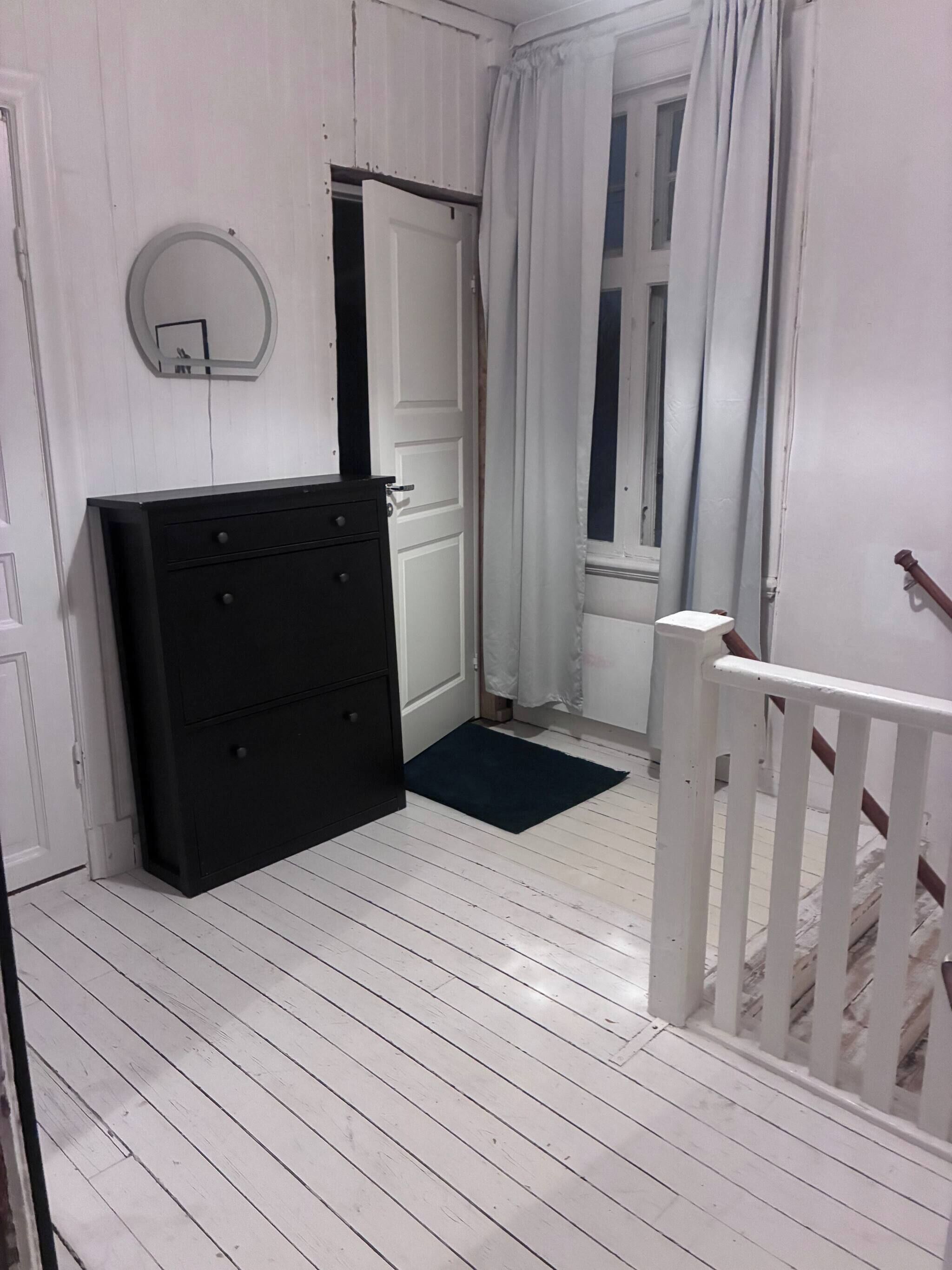 Passadís estret amb paviment de fusta blanca, un armari negre, una porta blanca i una finestra amb cortines blanques.