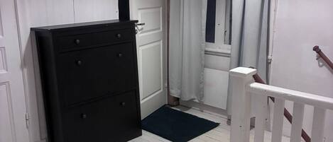 Passadís estret amb paviment de fusta blanca, un armari negre, una porta blanca i una finestra amb cortines blanques.