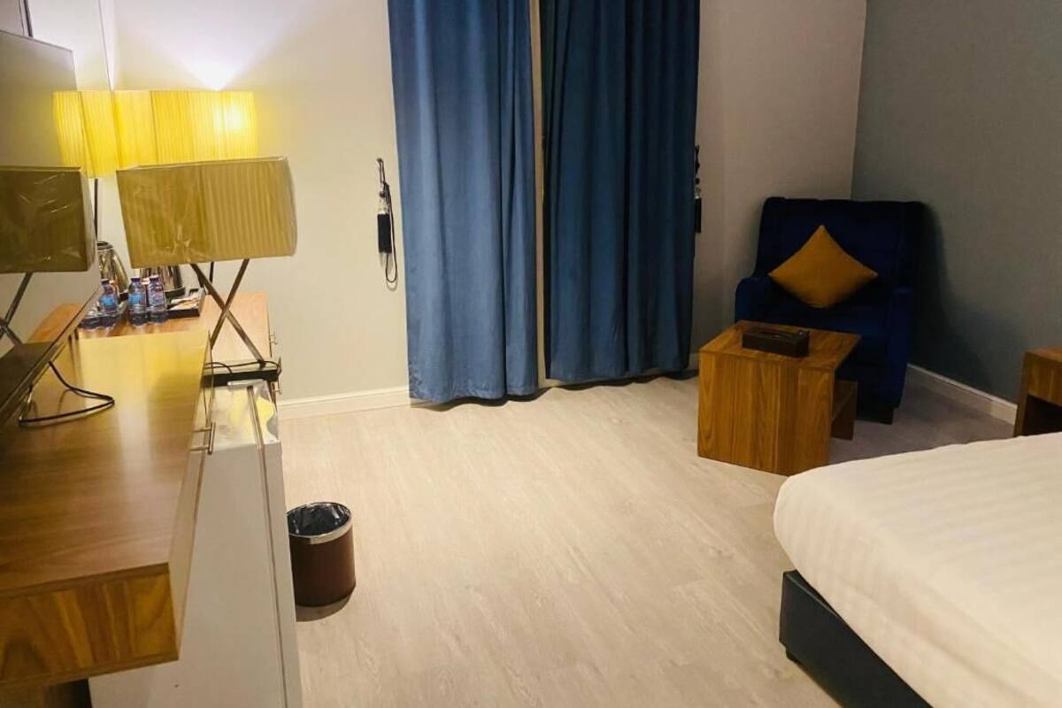 Una habitación de hotel con una cama, un escritorio con lámpara, una silla, una mesita y un cesto de basura.