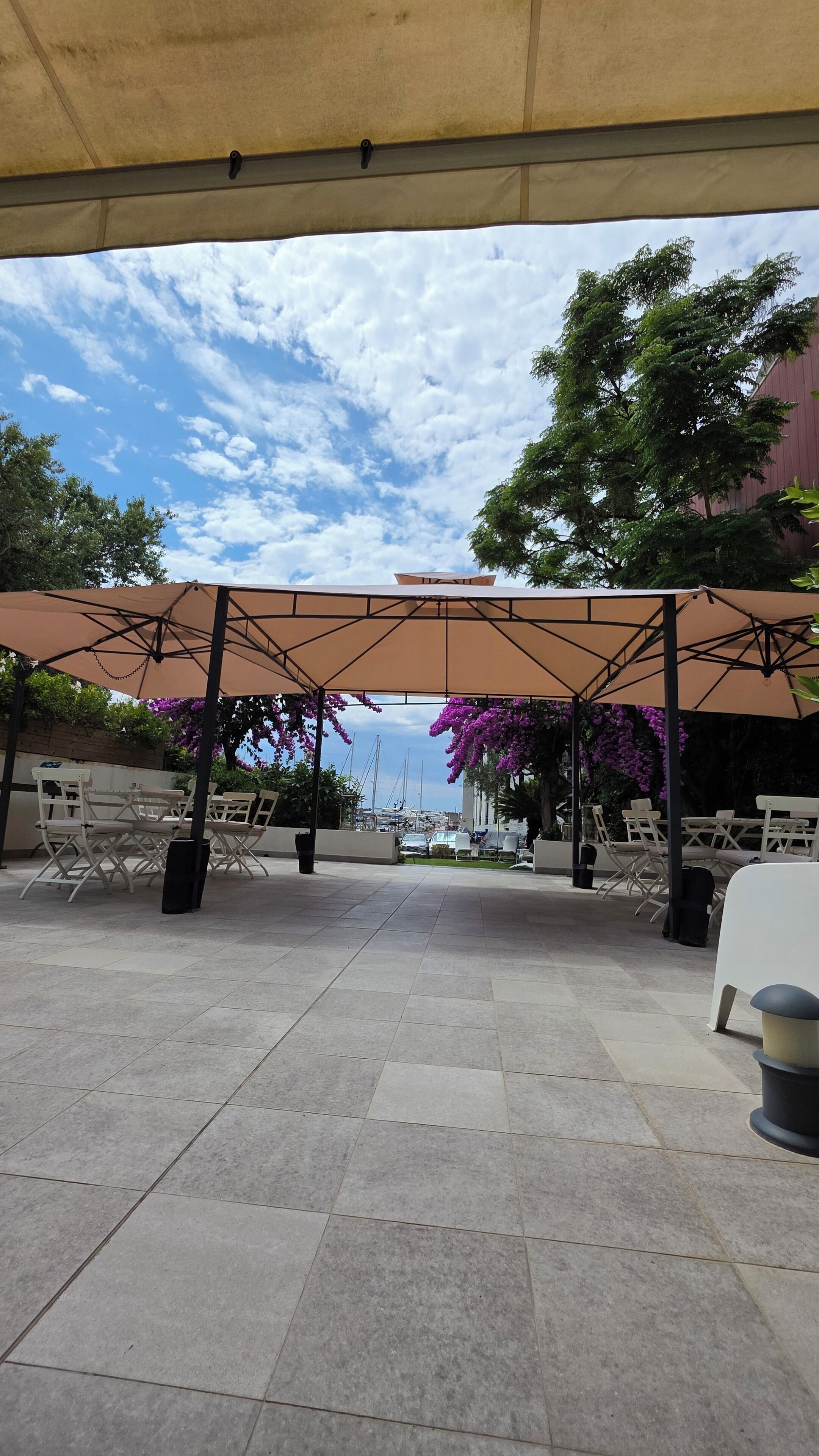 An outdoor patio area with tiled flooring, covered by a canopy, and furnished with white chairs and tables.
