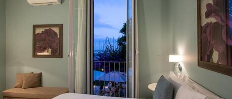 A hotel room with a bed, a window with a view of the outside, a wall-mounted air conditioner, and a framed picture on the wall.