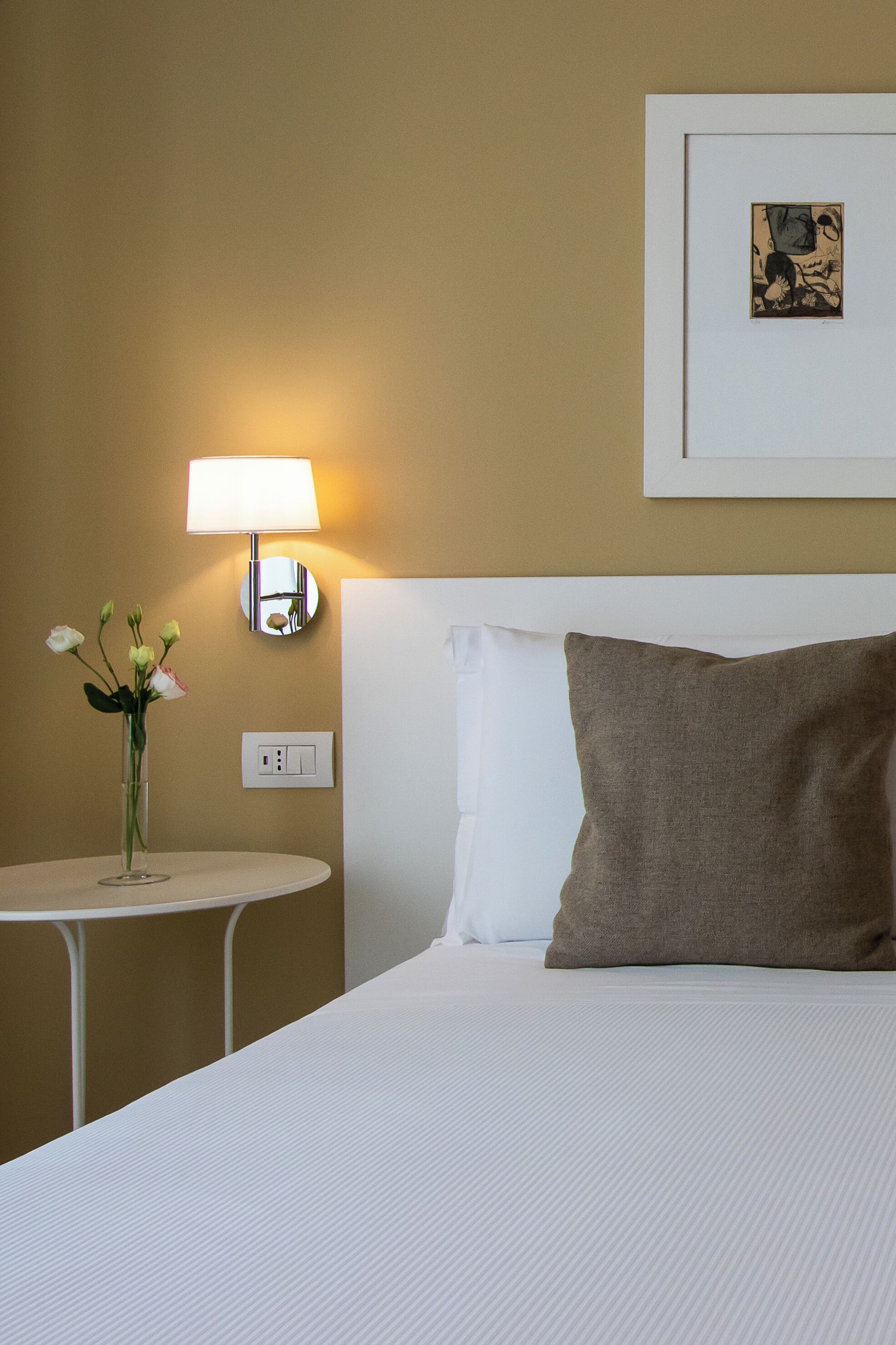 A hotel room with a bed, a bedside table with a lamp, a vase with flowers, and a framed picture on the wall.