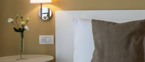A hotel room with a bed, a bedside table with a lamp, a vase with flowers, and a framed picture on the wall.