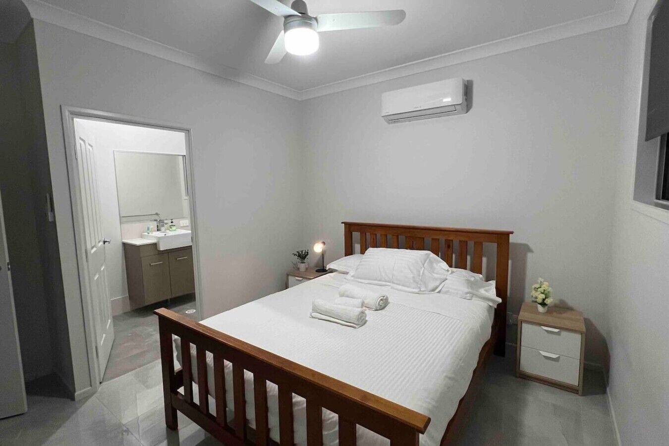 Quarto com cama de madeira, mesa de cabeceira, aparelho de ar condicionado e ventoinha de teto.