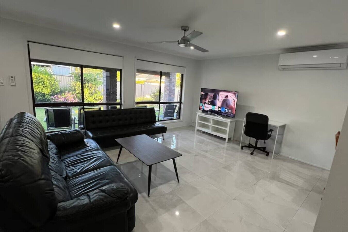 Uma sala de estar moderna com um sofá de couro preto, uma pequena mesa de centro, uma TV em um suporte branco e um ventilador de teto.