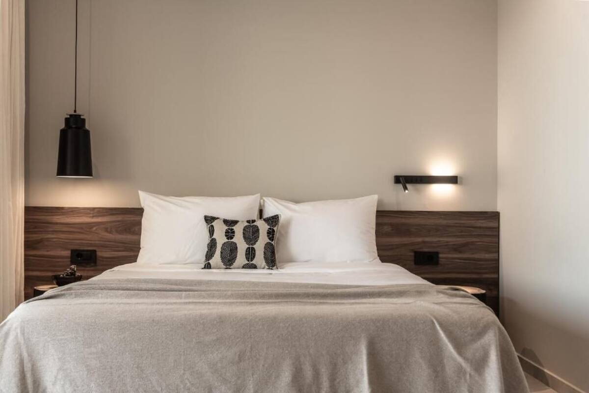 A neatly made bed with a grey bedspread and two pillows, one plain and one with a leaf pattern. There is a black pendant light hanging above the bed and a wall-mounted light on the right side of the headboard.