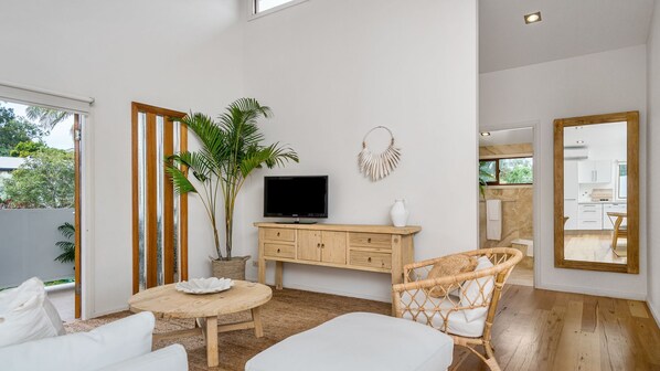 Una sala de estar moderna con televisor, muebles de madera, una planta grande y vista a la cocina.