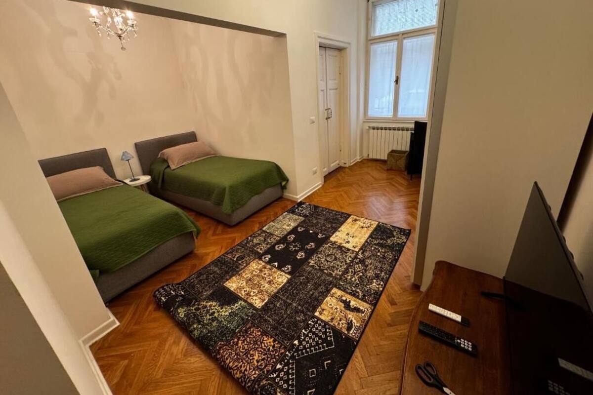 A small, well-lit room with two beds, a large patchwork rug, a chandelier, and a television.