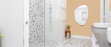 A modern bathroom with a glass shower enclosure, a small wooden stool with a plant, and a round mirror on the wall.