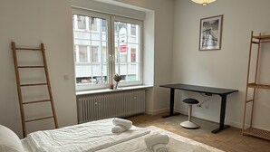 Un dormitorio con dos camas, un escritorio, una silla, una escalera, un radiador y una ventana con vistas a edificios.