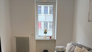 Un salón con un sofá gris, una mesita de centro de madera, una alfombra estampada, un radiador y una ventana con vistas a edificios.