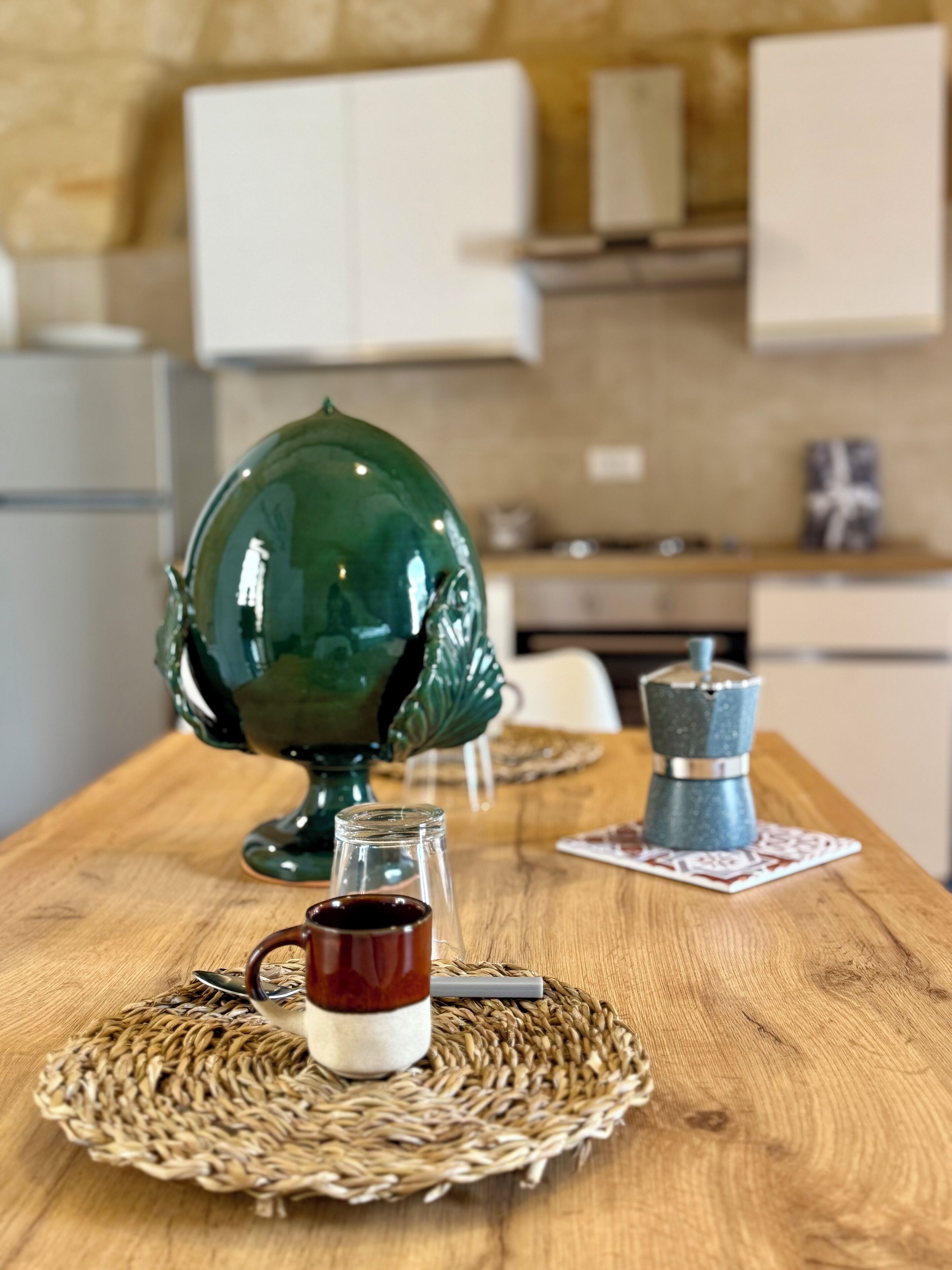 Una mesa de madera con un mantel tejido, una jarra de cerámica verde, un frasco de vidrio con líquido oscuro y una cafetera azul.