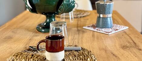 Una mesa de madera con un mantel tejido, una jarra de cerámica verde, un frasco de vidrio con líquido oscuro y una cafetera azul.