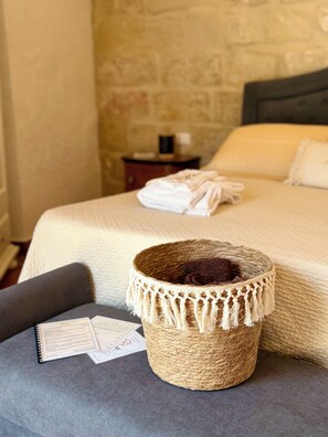 Una cama bien hecha con cabecera, una pequeña cesta redonda tejida con flecos, un sillón gris y una mesita de noche con cafetera.