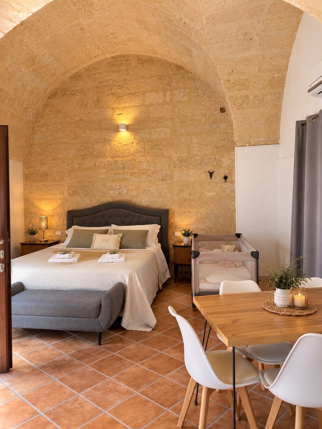 A bedroom with a stone wall, a bed with pillows, a bedside table with a lamp, and a chair.