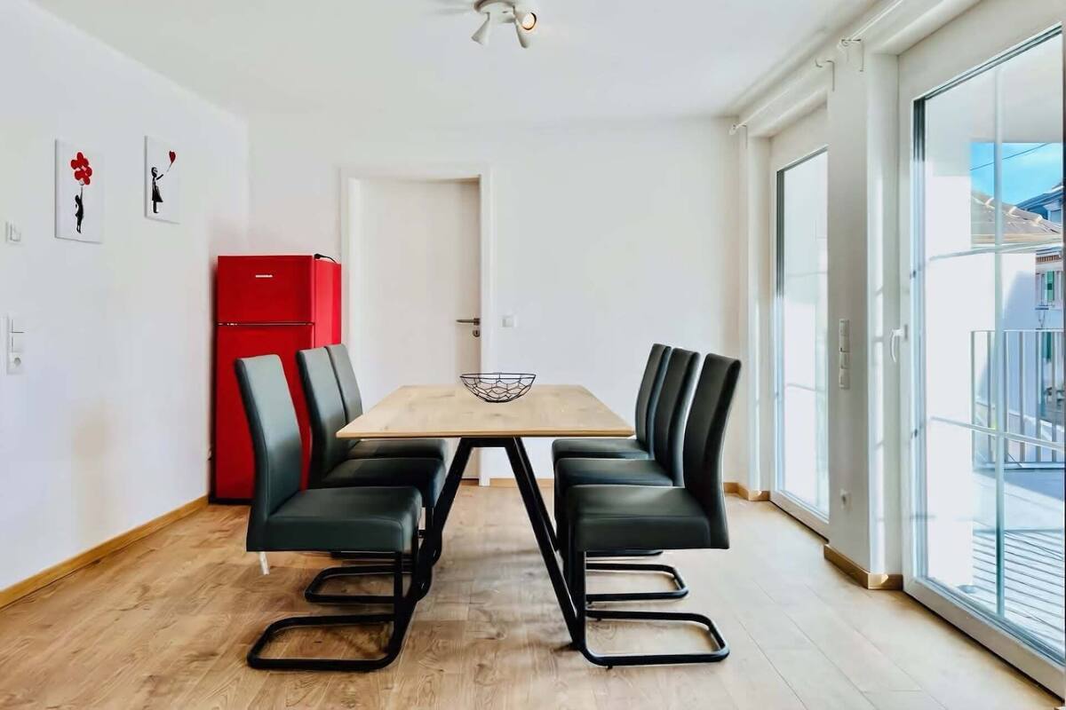 Ein moderner Essbereich mit einem Holztisch und schwarzen Stühlen, einem roten Kühlschrank und einem großen Fenster mit Blick nach draußen.
