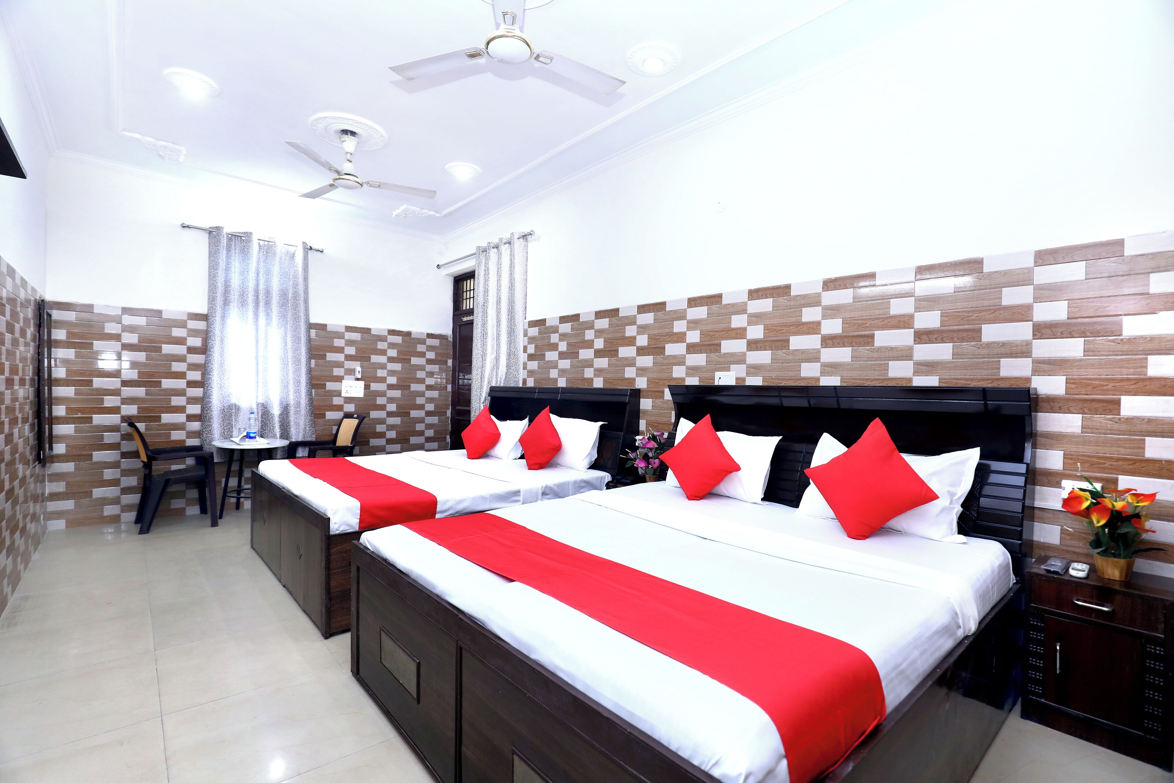 A hotel room with two beds, a ceiling fan, a small table with chairs, a nightstand, and a decorative vase.