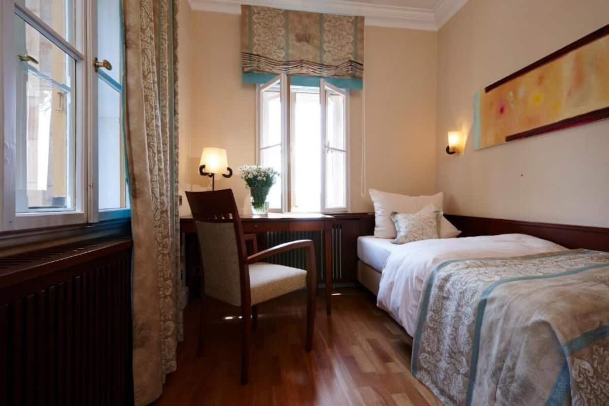 Et hotelværelse med seng, skrivebord med stol, en radiator og et vindue med gardiner.