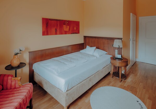 A neatly arranged hotel room with a bed, a small table with a lamp, a chair, and a round table. - LA VILLA am Starnberger See (Poecking)