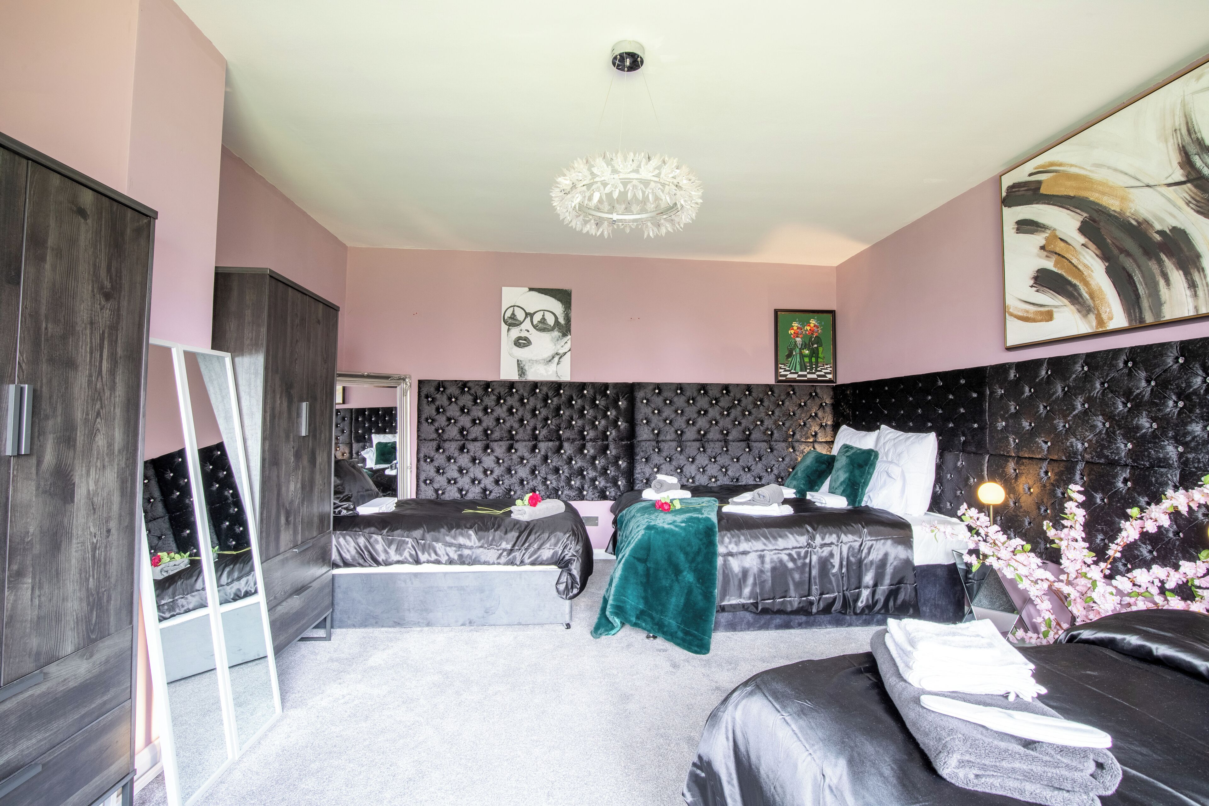 A modern bedroom with a large bed, a smaller bed, and a seating area. There is a mirror, a chandelier, and artwork on the walls.