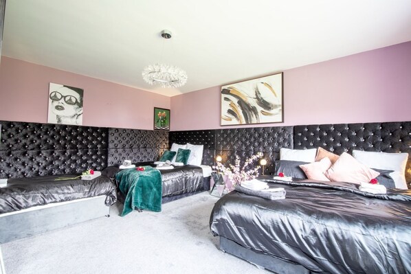 A modern bedroom with two large beds, a tufted headboard, a chandelier, and artwork on the walls. - Group Accommodation | 8 Single Beds | Private Gated Property | On-Site Parking (Rushden)