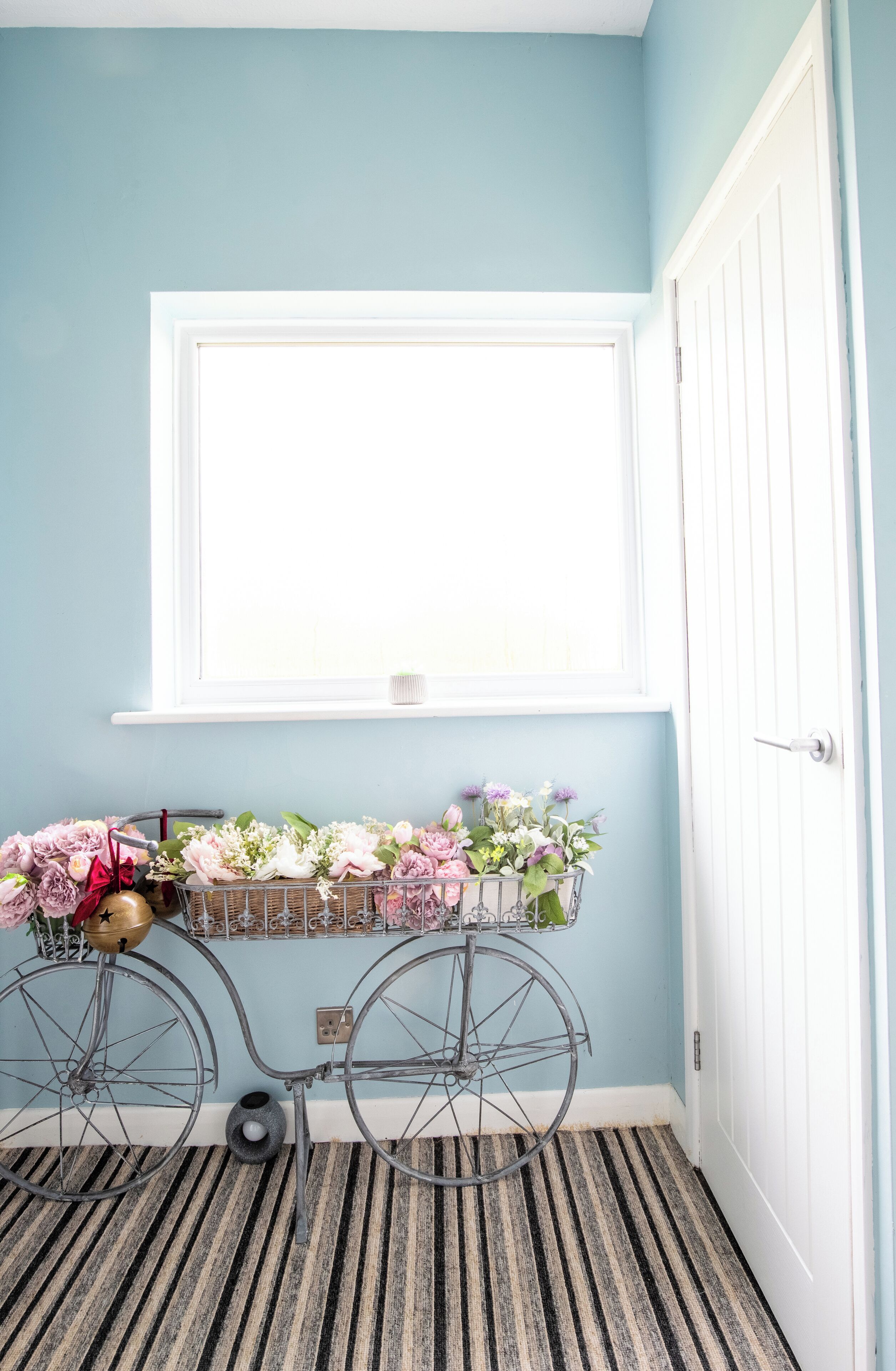 Een kamer met een raam, een fiets met een bloemenmand, en een gestreept tapijt.